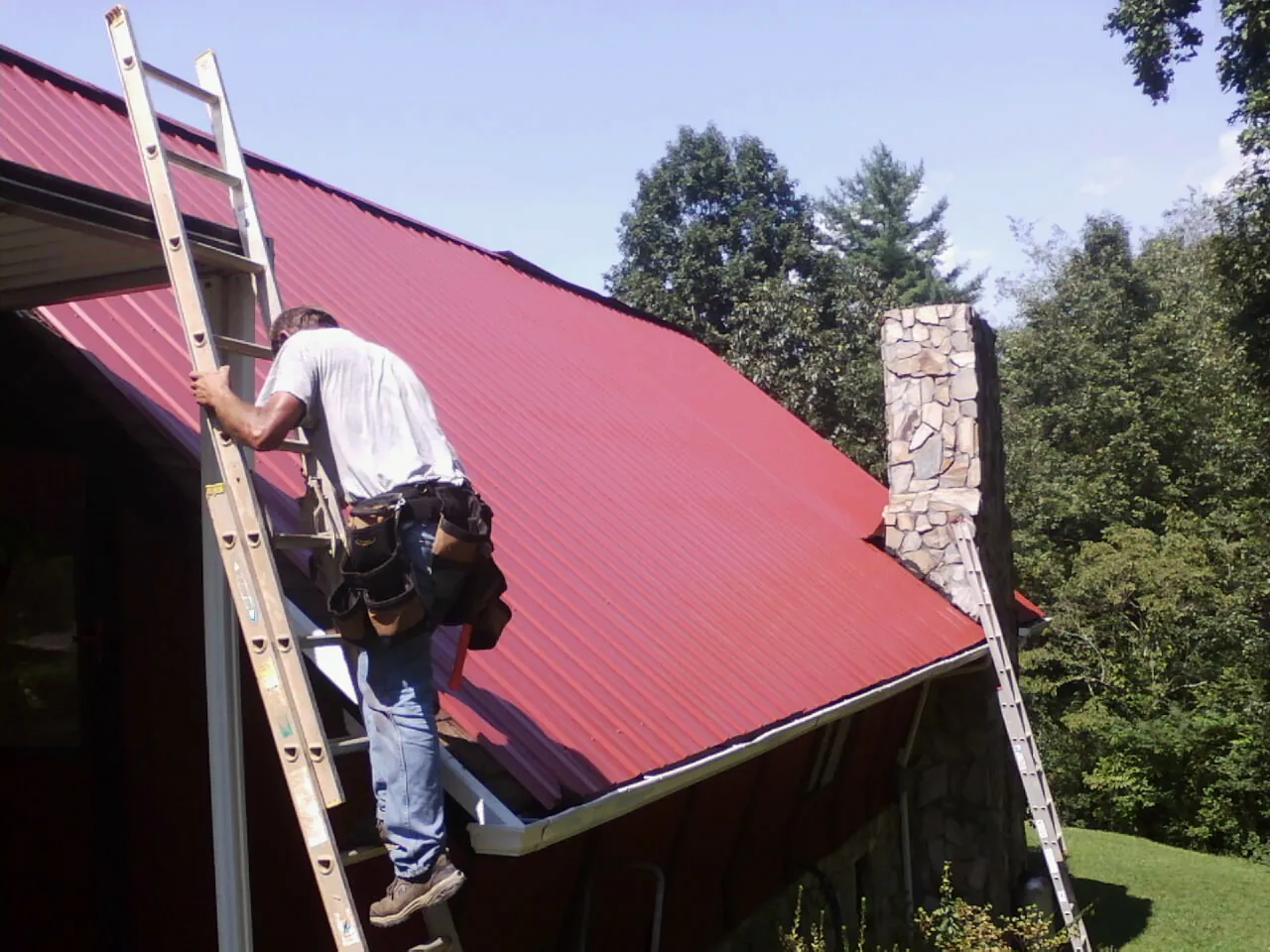 Roofer on a ladder beside a red metal roof and stone chimney — Soffit & Fascia Repair by Roofing Brothers in Yalaha